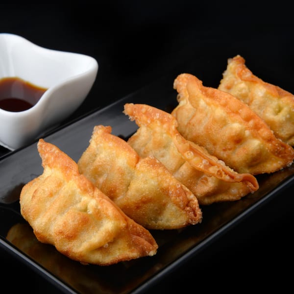 Five golden-fried gyoza dumplings on a black rectangular plate, with a white sauce dish beside them.