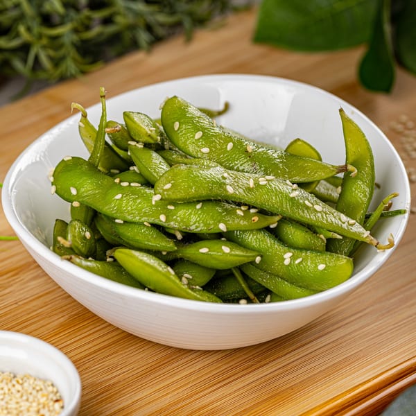 A white bowl filled with vibrant green edamame pods, sprinkled with sesame seeds, on a wooden surface.