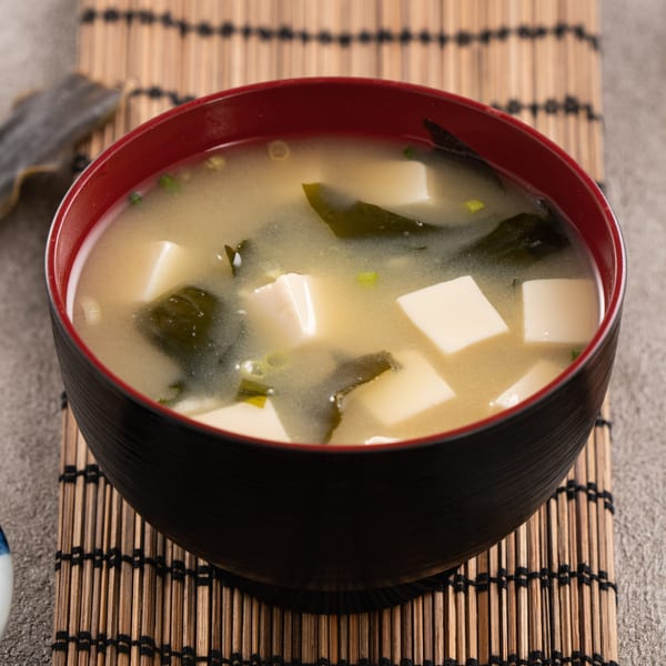 A black and red bowl of miso soup with tofu and seaweed, on a bamboo mat.